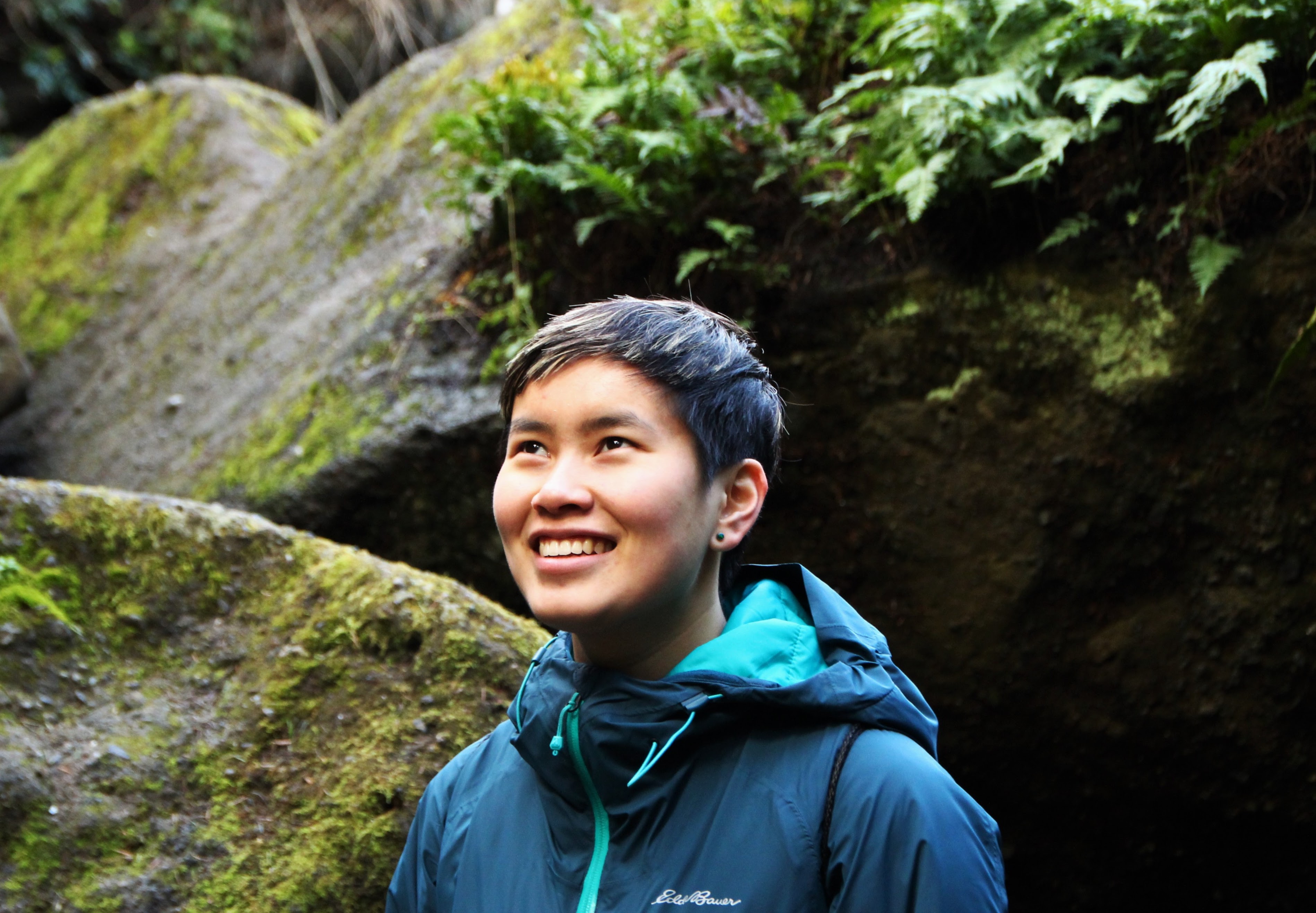Young asian person (Katie) smiling while looking upward in front of large mossy rocks